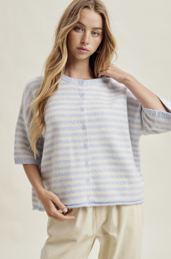 A woman modeling a light blue and white striped knit cardigan with a button-up front, paired with cream-colored pants, against a plain beige studio background.
