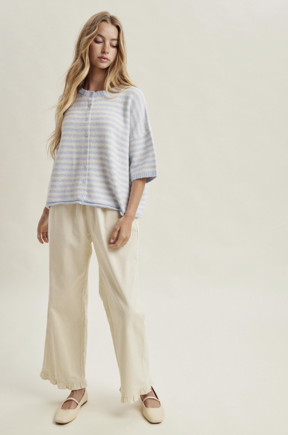 A woman modeling a light blue and white striped short-sleeve button-up shirt with cream wide-leg trousers and beige flat shoes, set against a plain cream studio background.