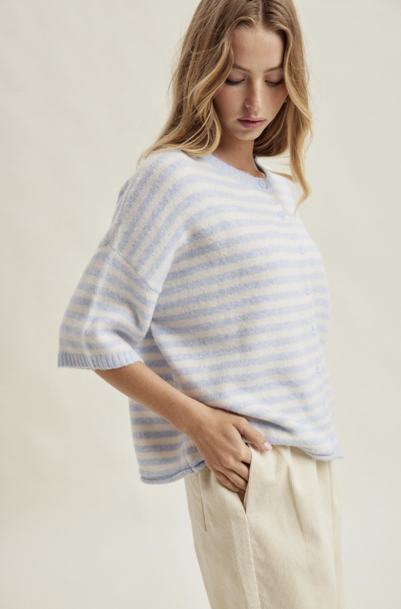 A woman wearing a light blue and white striped knit sweater with elbow-length sleeves, paired with cream-colored trousers, photographed against a plain beige studio background.