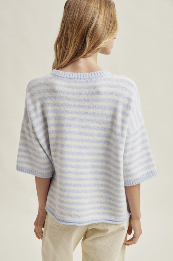 A woman shown from the back wearing a light blue and white horizontally striped knit sweater with elbow-length sleeves, paired with beige trousers, against a plain cream background.
