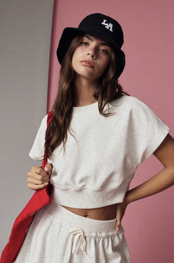 Woman wearing a white crop top and gray sweatpants with a red jacket, standing against a pink background.