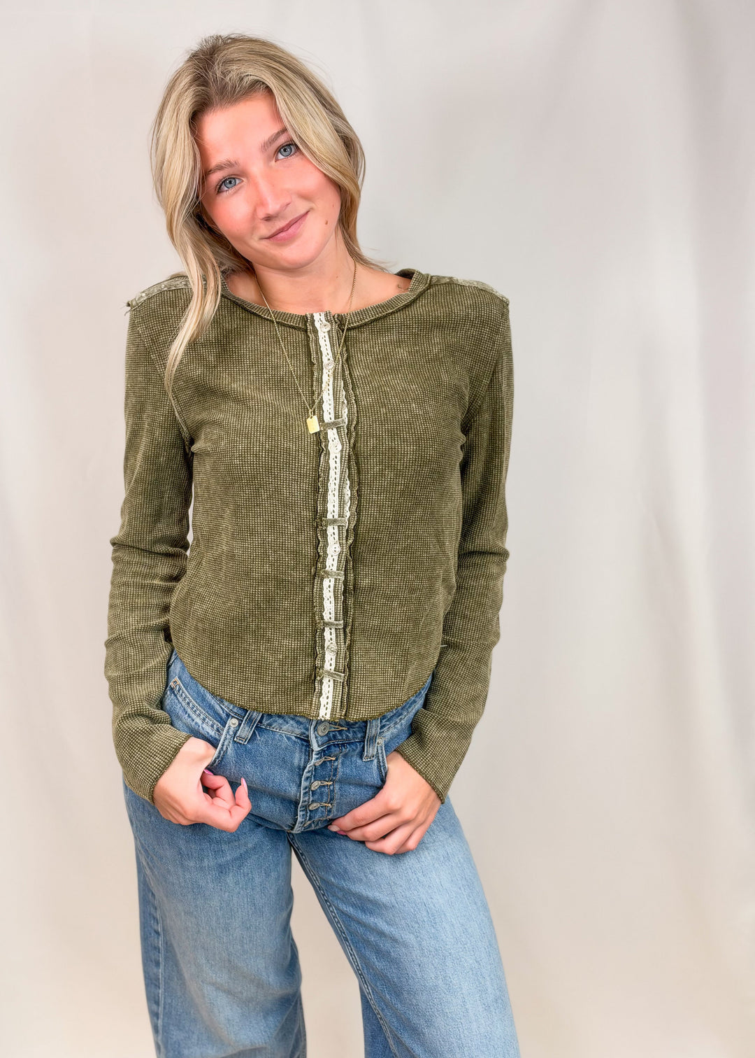 A woman modeling a cropped, long-sleeve olive green knit top with a decorative vertical cream and green patterned placket, paired with blue jeans, against a neutral studio backdrop.