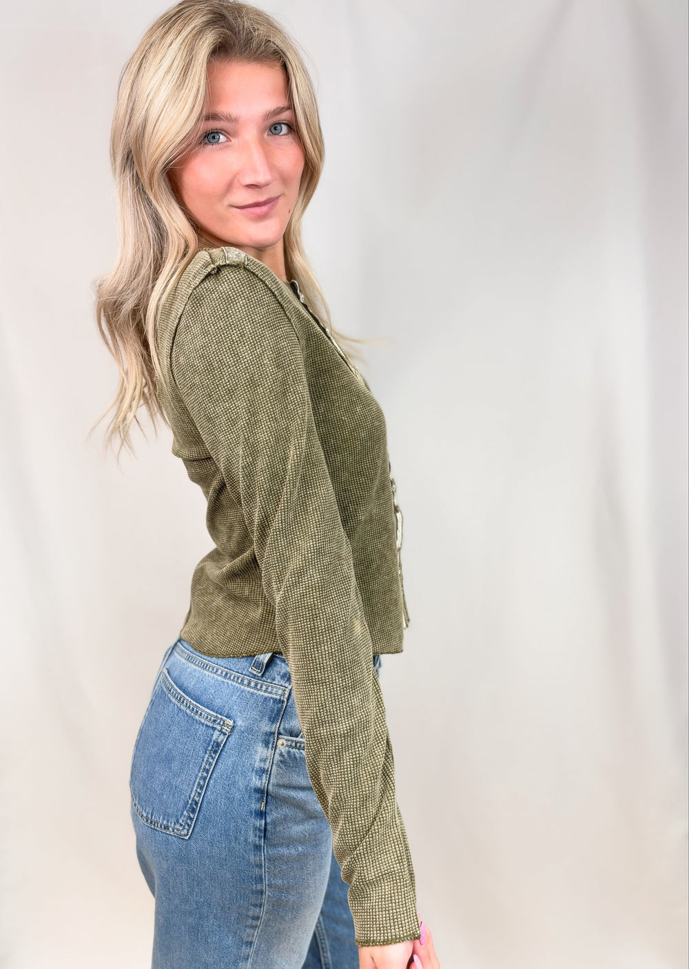 A woman with blonde hair wearing an olive green ribbed knit cardigan and blue jeans, posing in profile against a white studio backdrop.