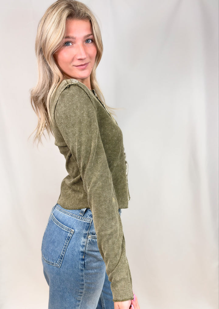 A woman with blonde hair wearing an olive green ribbed knit cardigan and blue jeans, posing in profile against a white studio backdrop.