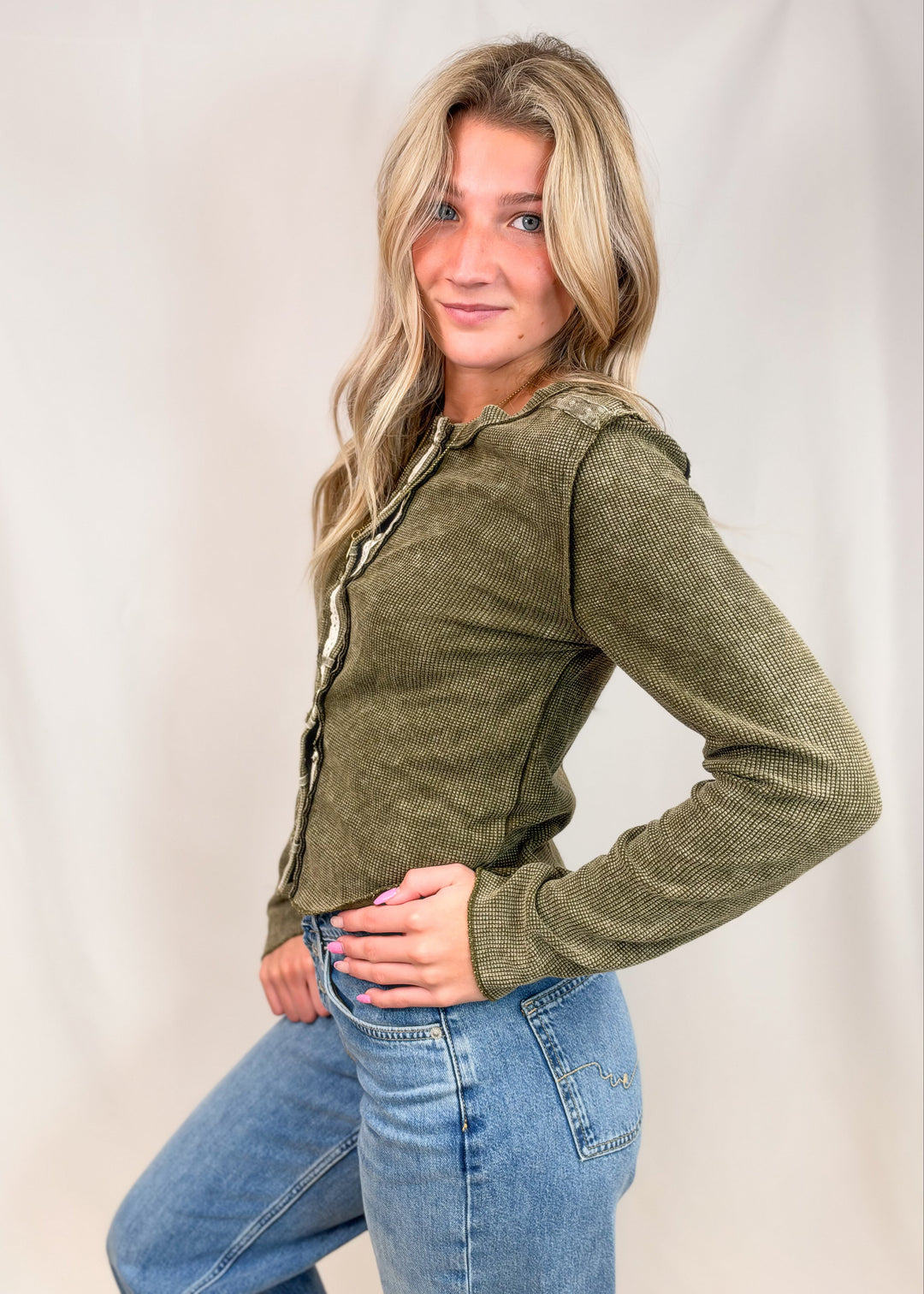 A woman modeling a textured olive green long-sleeve top with a decorative lace-up front detail, paired with blue jeans, against a neutral studio backdrop.