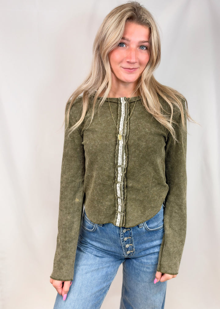A woman with blonde hair wearing an olive green long-sleeve top with a decorative cream and green patterned vertical placket, paired with blue jeans, standing against a light gray studio backdrop.