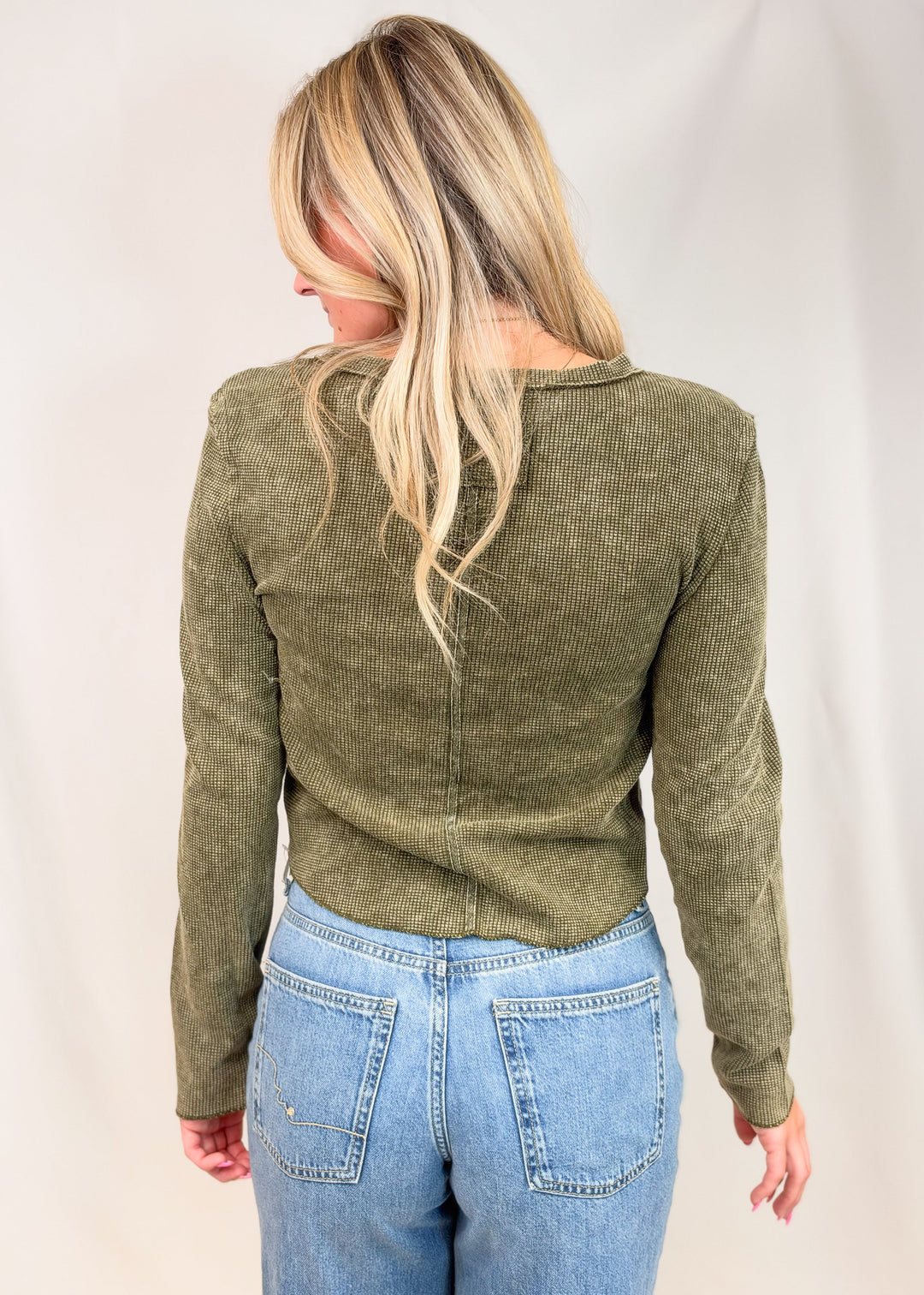 A woman shown from the back wearing an olive green long-sleeve ribbed knit top and light blue jeans, standing against a plain light background.