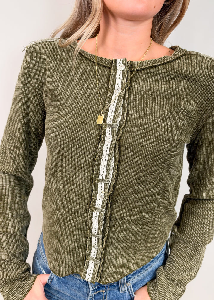 An olive green ribbed knit long-sleeve top with a white lace trim running down the front, worn by a model with blue jeans.