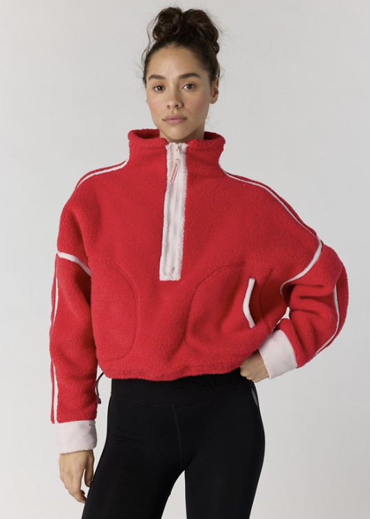 A woman modeling a red cropped fleece pullover with a white half-zip, white sleeve stripes, and white ribbed cuffs, paired with black leggings, against a light gray studio background.