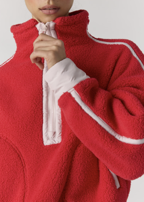 A close-up of a person wearing a red sherpa fleece quarter-zip pullover with white contrast piping and a white inner zipper pull, shown against a light gray studio background.