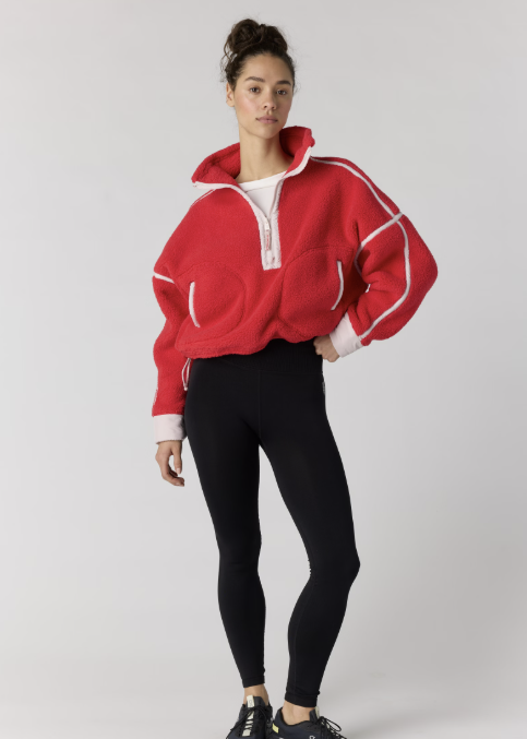 A woman modeling a red fuzzy half-zip pullover sweatshirt with white contrast stitching and a white inner collar, paired with black leggings and black sneakers, against a light gray studio background.