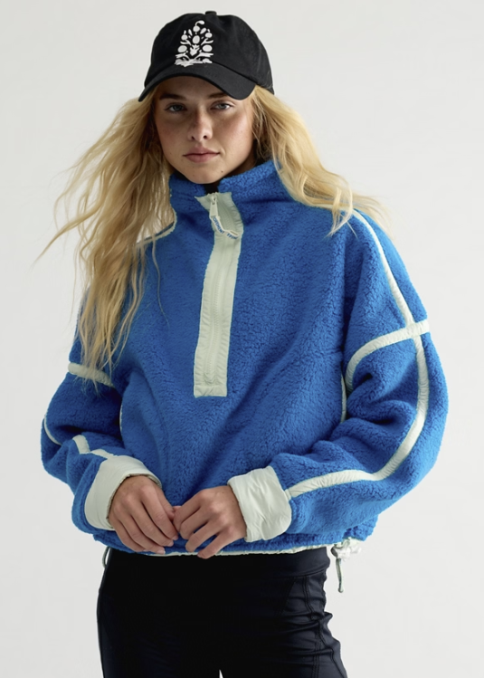 Woman wearing blue fleece jacket with mint details and black pants and baseball cap on plain background
