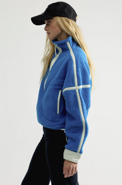 Woman wearing blue fleece jacket with mint details and black pants and baseball cap on plain background
