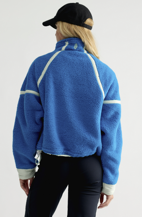 Woman shown from back wearing blue fleece jacket with mint details and black pants and hat on plain background