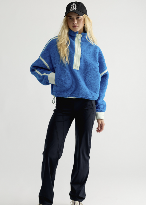 Woman wearing blue fleece jacket with mint details, black pants and black baseball cap on plain background