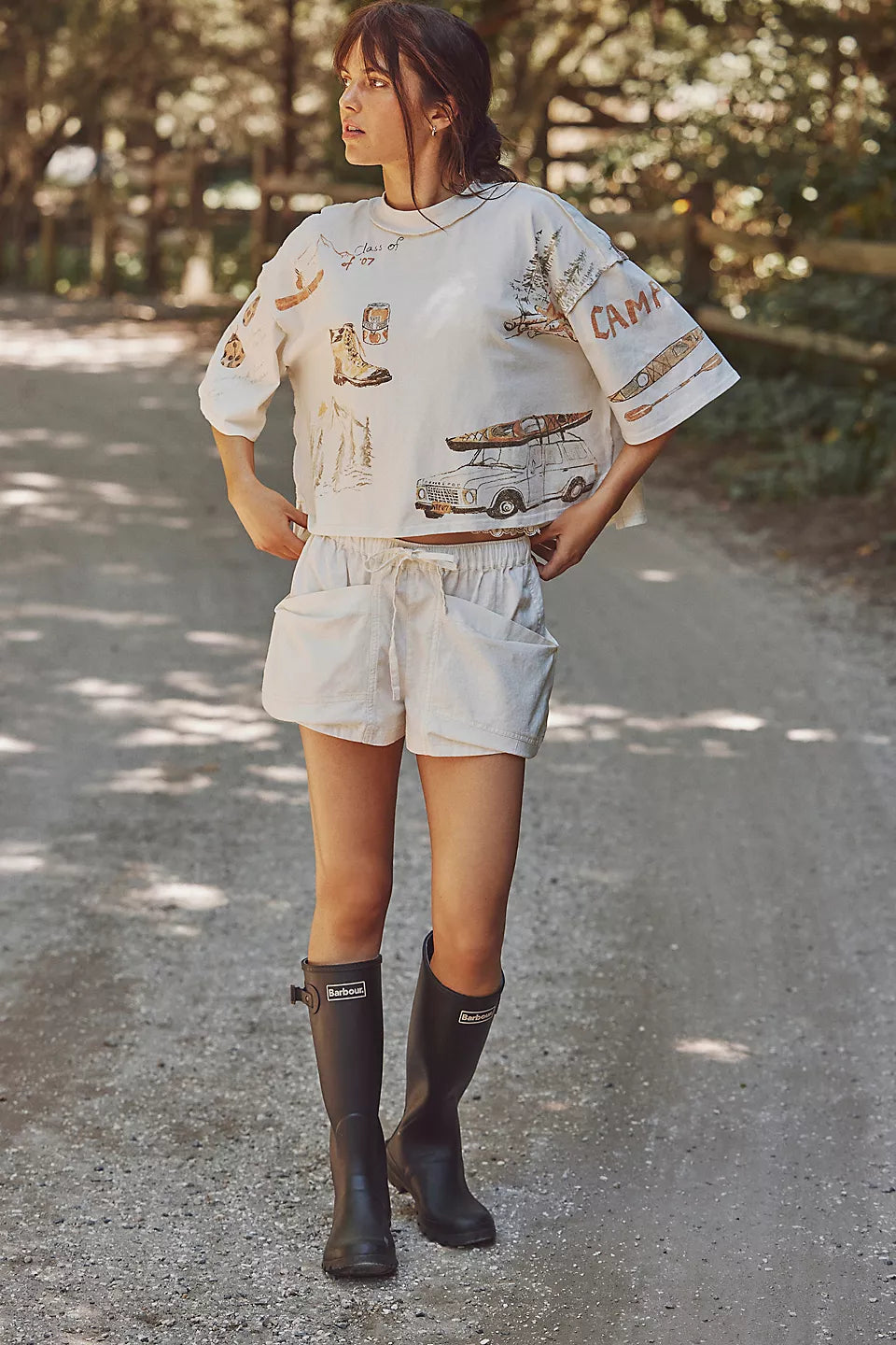 Woman wearing a white graphic t-shirt, white shorts, and black rain boots on a road with trees in the background.