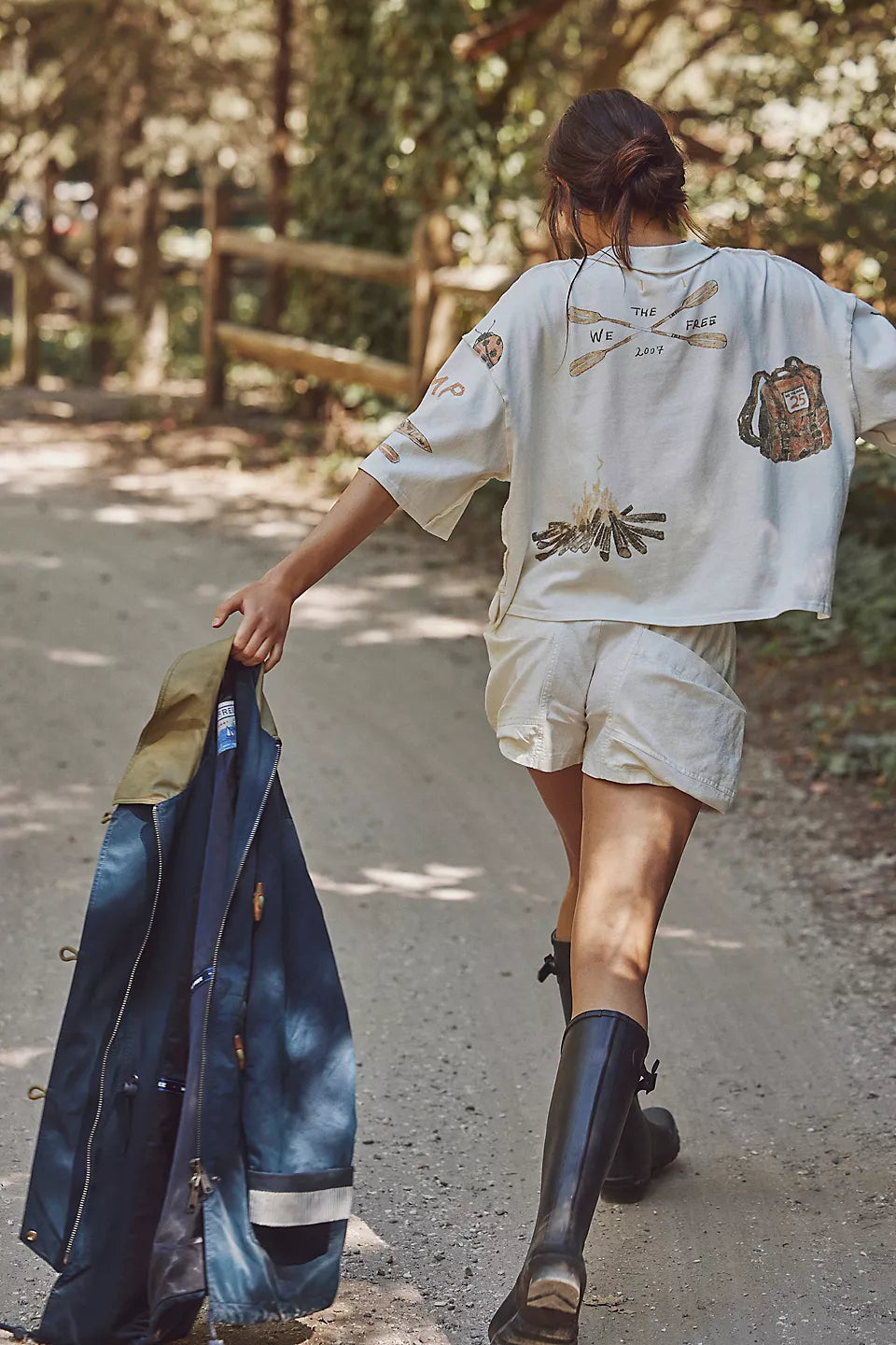 Person walking outdoors holding a blue jacket, wearing a white shirt with patches and beige shorts.