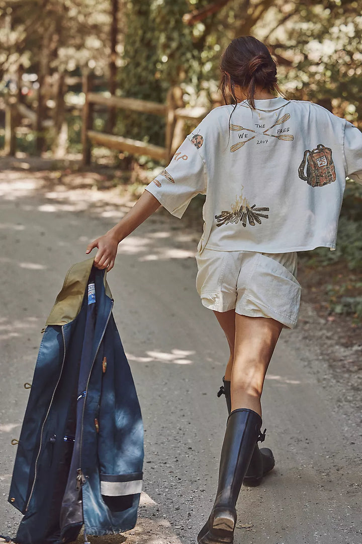 Person walking outdoors holding a blue jacket, wearing a white shirt with patches and beige shorts.