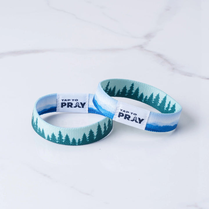 Two wristbands with a pine tree landscape design in shades of blue, teal, and white, featuring a white paper band that reads &