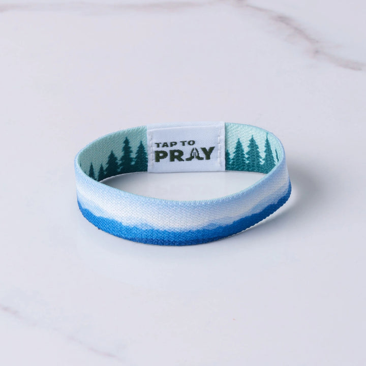 A fabric wristband with a blue and white mountain landscape design, green pine trees, and a white center tab that reads &