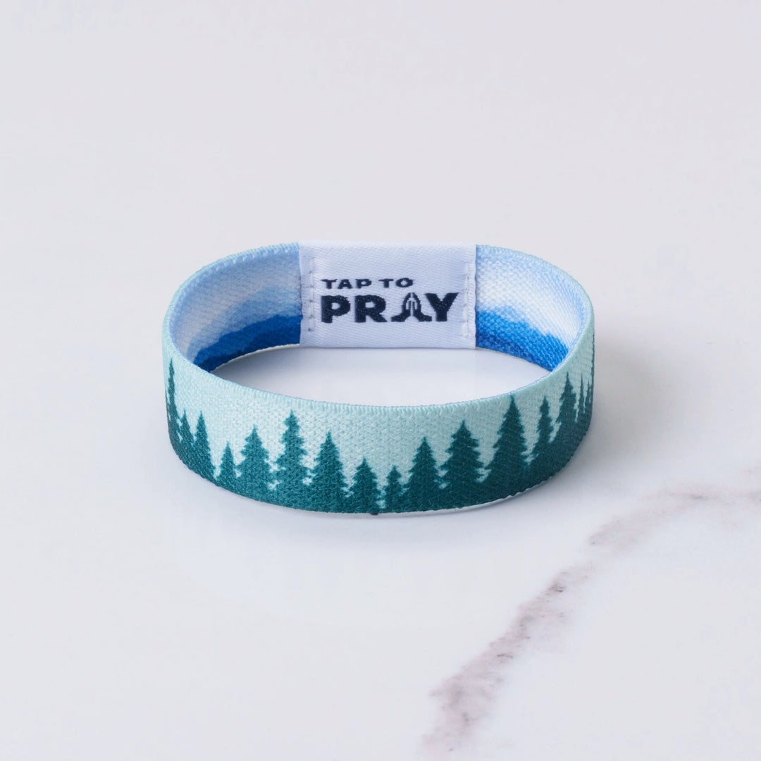 A fabric wristband with a pine tree pattern in shades of blue and teal, featuring a white inner band with the text &