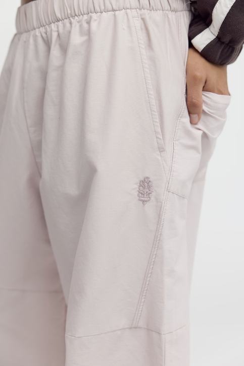Close-up of a person wearing light blush pink wide-leg pants with an elastic waistband, side pockets, and a small embroidered logo on the thigh.