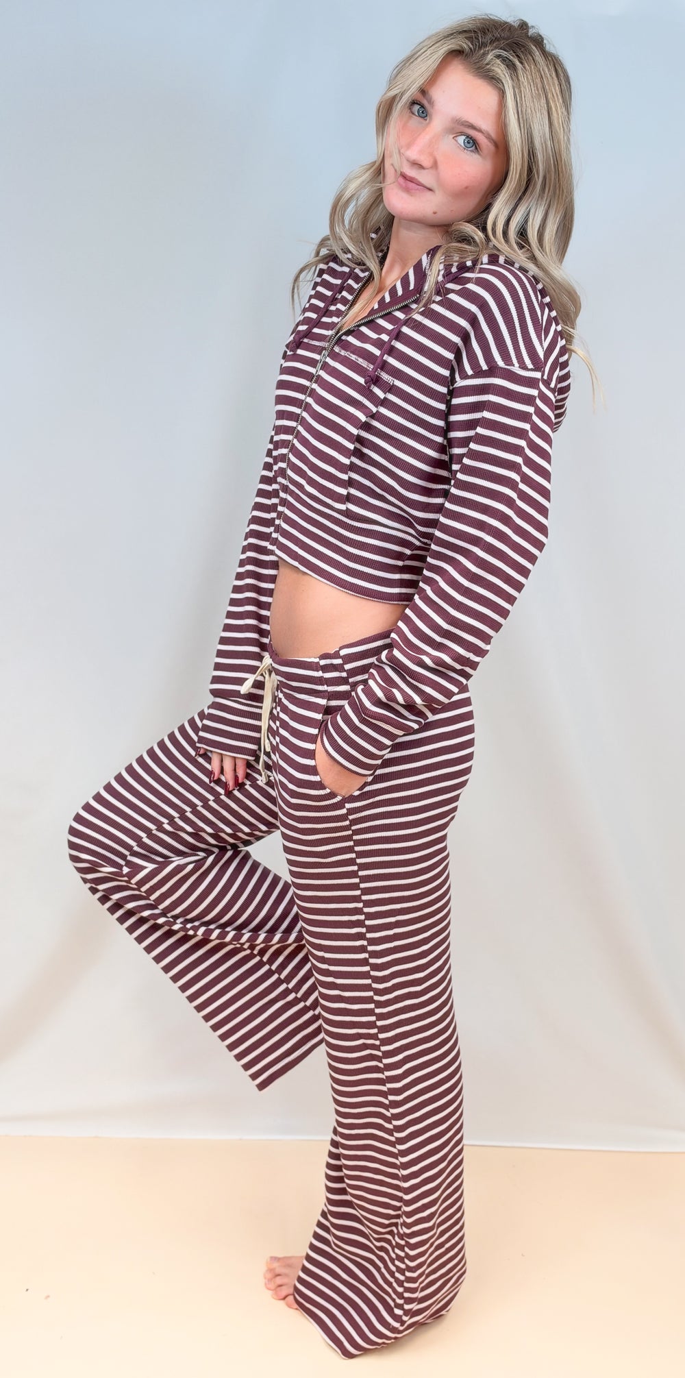 Woman from side view wearing matching burgundy and cream stripe set with zipper hoodie and pants on plain background