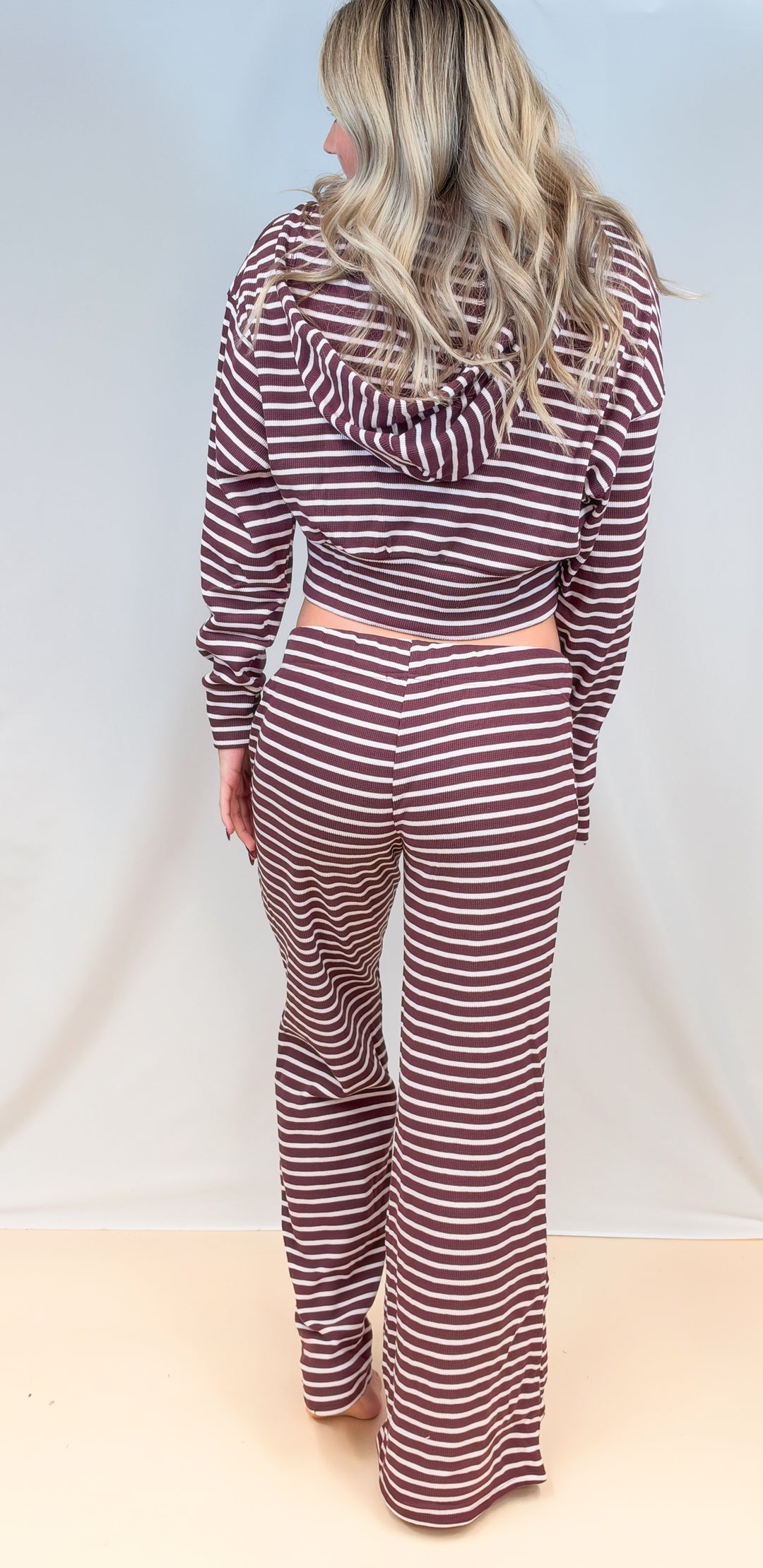Woman shown from back view wearing burgundy and cream stripe matching set with hoodie and pants on plain background