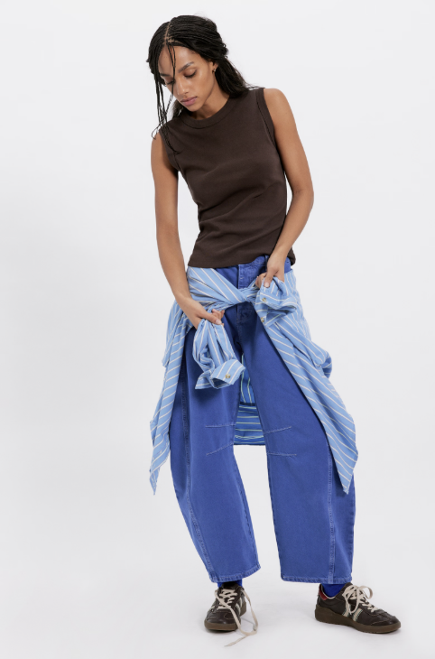 Woman wearing a brown sleeveless top and blue wide-leg pants holding a blue jacket on a white background