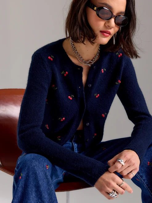 A woman seated on a brown leather chair wearing a navy blue fuzzy knit cardigan with small embroidered cherry motifs, blue jeans, layered silver chain necklaces, and silver rings. She is wearing dark sunglasses and looking downward against a plain light gray background.