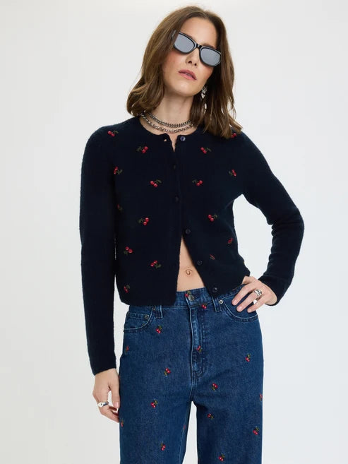A woman modeling a cropped navy cardigan with small red and green cherry embroidery, paired with blue jeans featuring matching cherry embroidery, against a plain light gray background.