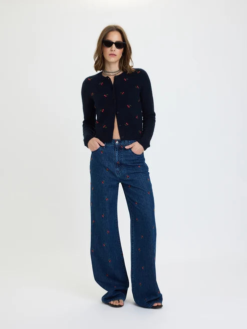 A model stands against a plain white studio background wearing a dark cropped cardigan and wide-leg blue jeans, both with small red embroidered motifs, along with black sandals and sunglasses.