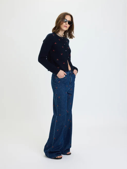 A woman modeling a dark navy blue knit cardigan with small red embroidered motifs, paired with wide-leg blue jeans featuring similar red embroidery, against a plain white studio background.