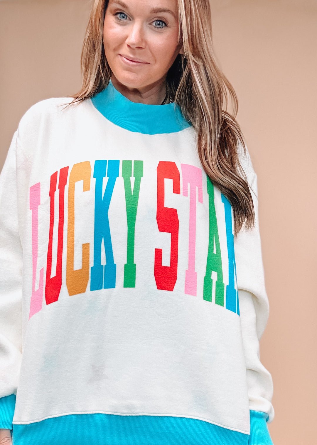 A woman wearing a cream-colored crewneck sweatshirt with a bright turquoise ribbed collar, cuffs, and hem, featuring the words &