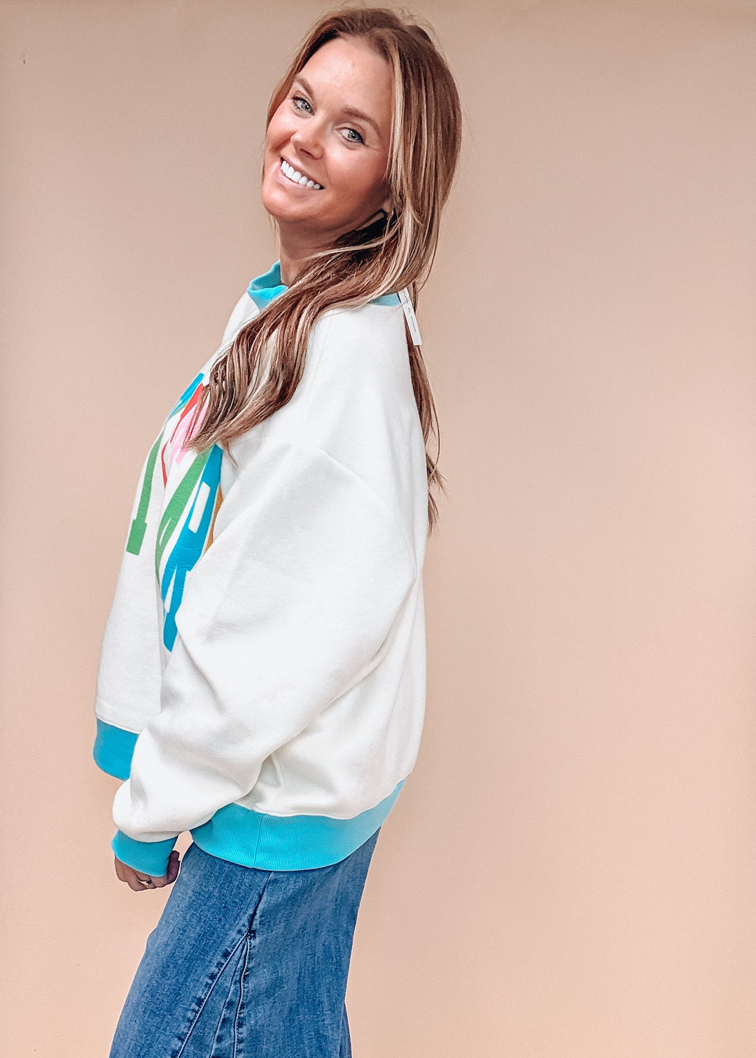 A woman modeling a white crewneck sweatshirt with bright turquoise ribbed trim and a colorful graphic on the front, paired with wide-leg blue jeans against a solid beige studio backdrop.