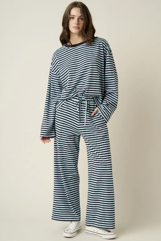 A woman modeling a matching two-piece loungewear set featuring a navy and white horizontal striped long-sleeve top and wide-leg pants, styled with white sneakers against a plain beige studio background.