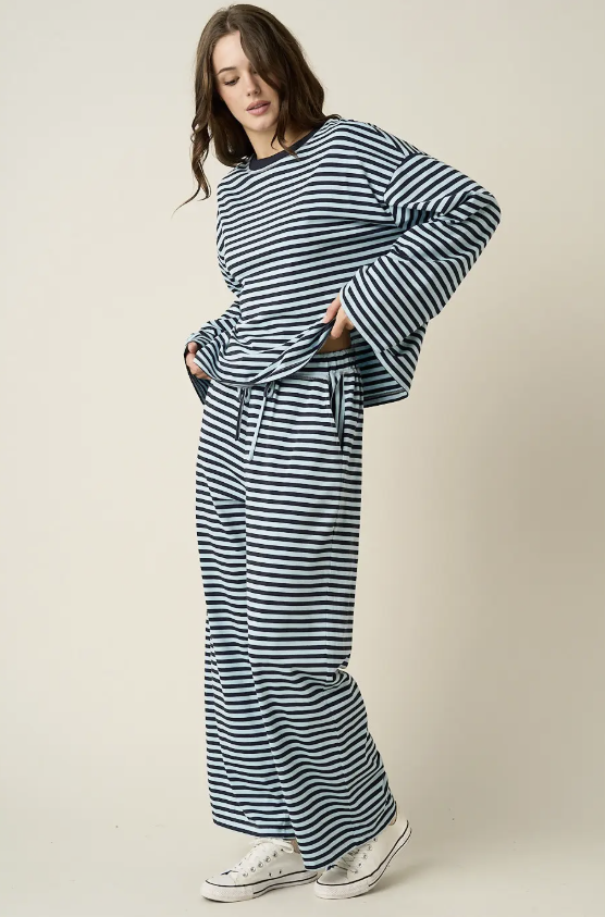 A woman modeling a matching navy and white striped lounge set, featuring a long-sleeve top with wide bell sleeves and matching wide-leg pants, styled with white canvas sneakers against a plain beige studio background.