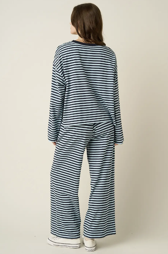 A model viewed from the back wearing a matching navy and white striped lounge set, consisting of a long-sleeve top and wide-leg pants, paired with white sneakers against a plain beige background.
