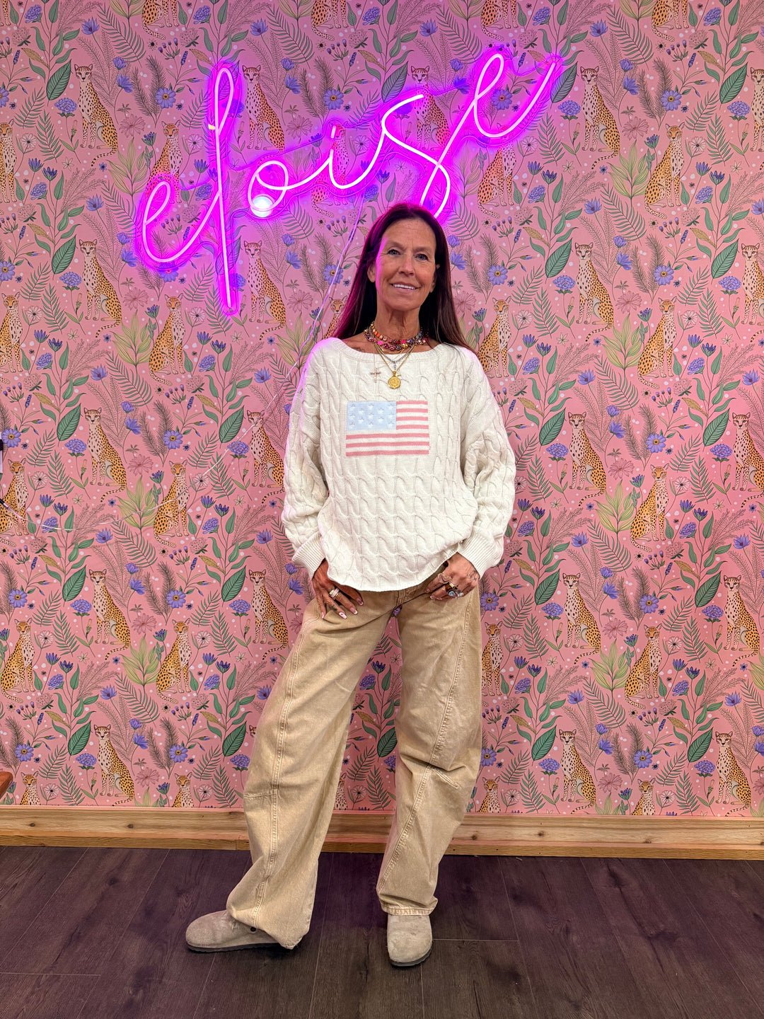 Woman wearing oatmeal cable knit sweater with american flag and beige pants on floral background