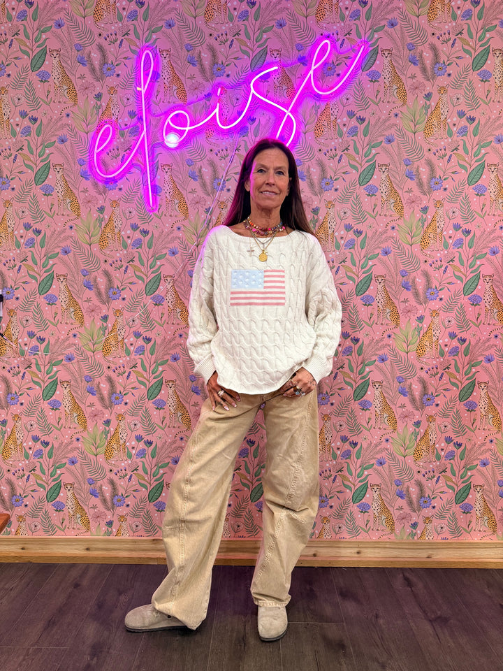 Woman wearing cream sweater with American flag and beige pants on floral background