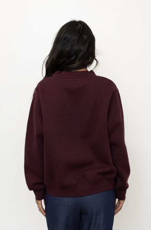 Woman shown from behind wearing a maroon mock neck pullover and jeans on white background