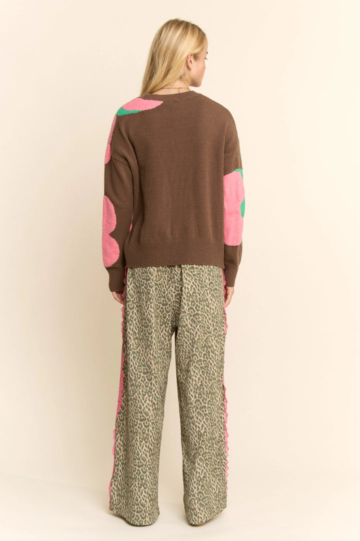 Woman wearing a brown sweater with pink and green accents and patterned pants on a beige background