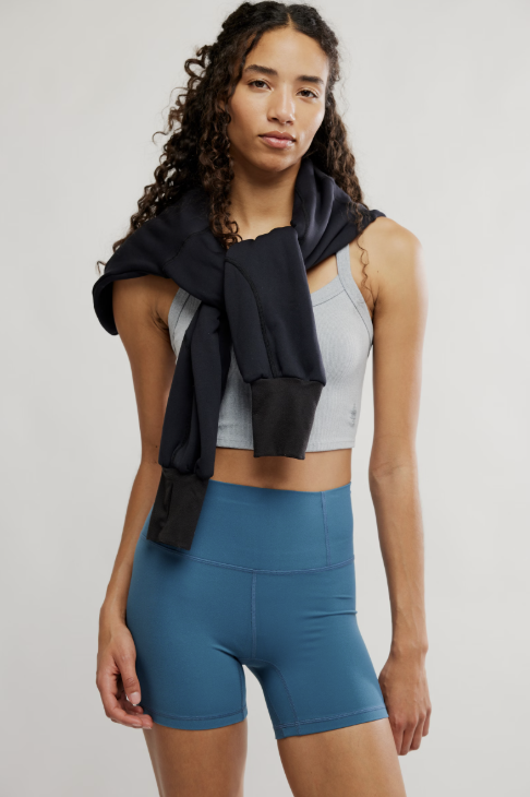 Woman wearing a gray tank top and blue shorts with a black towel over her shoulder on a light background