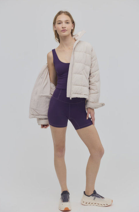 Woman wearing purple cami and bike shorts with cream color puffer jacket on plain background