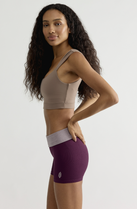 A woman modeling a two-piece activewear set consisting of a taupe sports bra and textured purple athletic shorts with a light gray waistband, set against a plain light gray background.