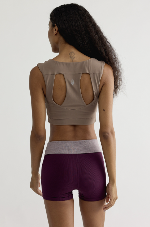 A woman shown from the back wearing a taupe-beige sports bra with large cutout details and dark purple ribbed athletic shorts against a plain light gray studio background.