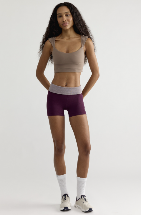 A woman modeling a matching activewear set consisting of a beige scoop-neck crop top and dark purple athletic shorts with a light gray waistband, paired with white socks and sneakers, against a plain light gray background.