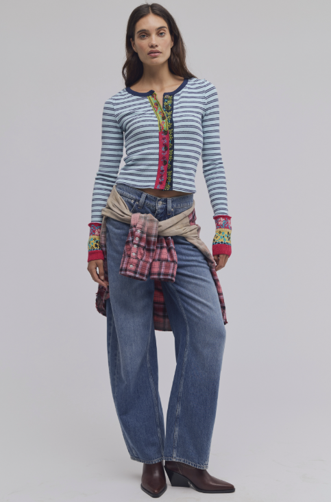 A model stands against a light gray studio background wearing a blue and white striped long-sleeve top with a colorful embroidered placket, blue jeans, a beige shirt tied around the waist, and brown ankle boots.