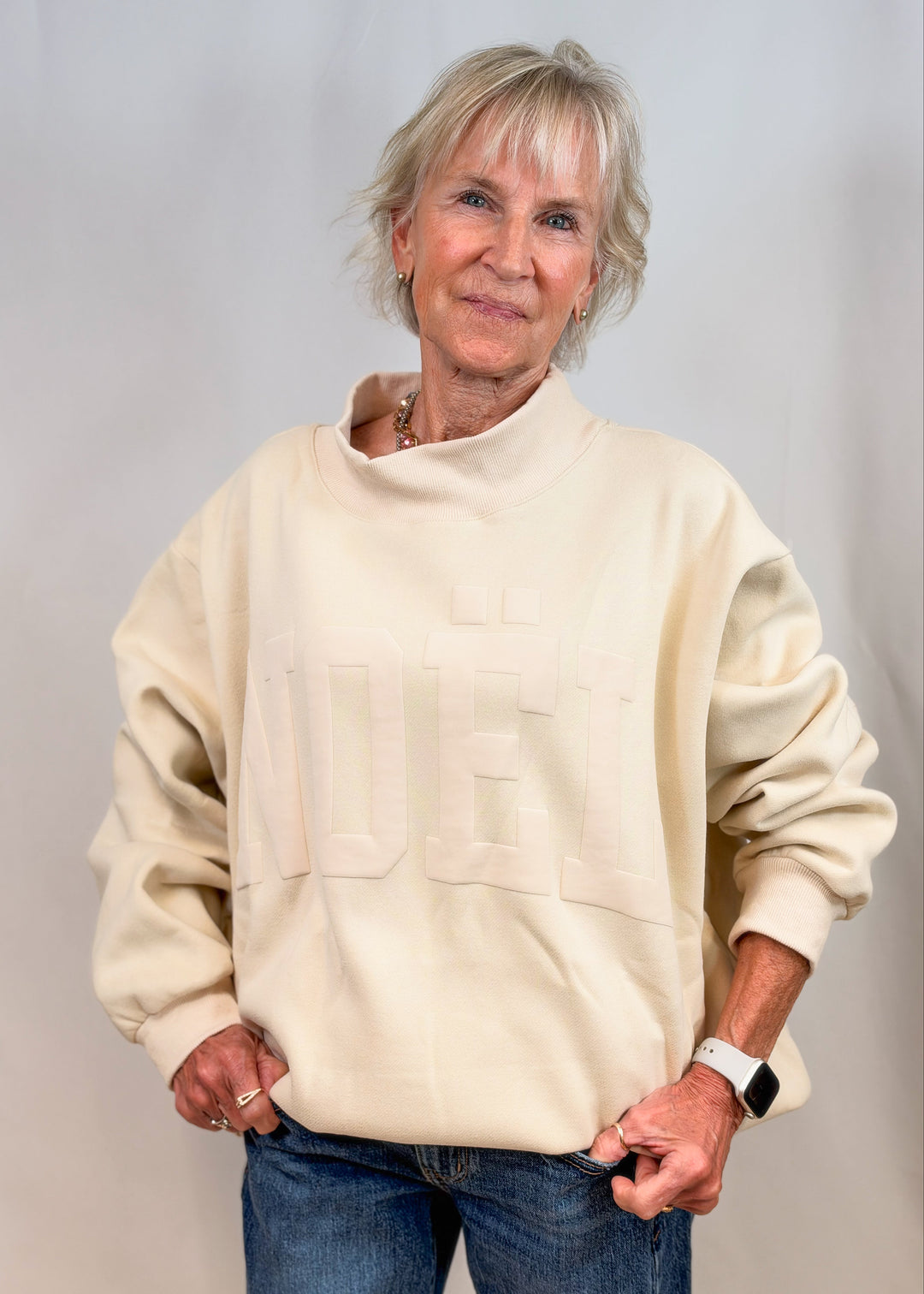 Woman wearing cream color mock neck swewatshirt with jeans on plain background. Sweatshirt is embossed with &