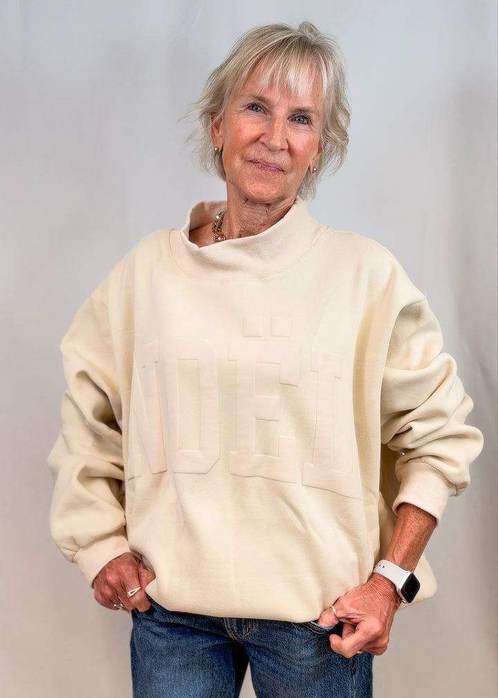 Woman wearing cream color mock neck swewatshirt with jeans on plain background. Sweatshirt is embossed with &